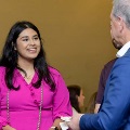 Two individuals stand indoors conversing, one wearing a bright pink outfit with honor cords and holding a card, facing another in a blue jacket, with other attendees and a neutral wall in the background.
