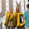 Two individuals hold large gold “M” and “D” balloons in front of illuminated “UTMB 24” letters while another person in the foreground takes a photo, with balloon clusters and a curtain backdrop visible.