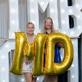 Two individuals stand indoors holding large gold “M” and “D” balloons in front of illuminated “UTMB 24” letters, with decorative red, white, gold, and silver balloon clusters beside a curtain backdrop.