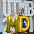 Two individuals stand indoors holding large gold “M” and “D” balloons in front of illuminated “UTMB 24” letters, with red, white, gold, and silver balloon clusters arranged on both sides against a curtain backdrop.