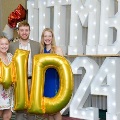 Three individuals stand indoors holding large gold “M” and “D” balloons in front of illuminated “UTMB 24” letters, with red, white, gold, and silver balloon clusters arranged on both sides against a curtain backdrop.