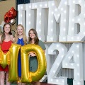 Three individuals stand indoors holding large gold “M” and “D” balloons in front of illuminated “UTMB 24” letters, with red, white, gold, and silver balloon clusters on both sides against a curtain backdrop.
