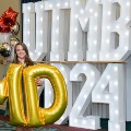 Individual stands indoors holding large gold “M” and “D” balloons in front of illuminated “UTMB 24” letters, flanked by red, white, gold, and silver balloon clusters against a curtain backdrop.