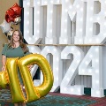 Individual standing indoors holding large gold “M” and “D” balloons in front of illuminated “UTMB 24” letters, with red, white, gold, and silver balloon clusters arranged on both sides against a curtain backdrop.