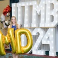 Individual in a blue dress stands indoors holding large gold “M” and “D” balloons in front of illuminated “UTMB 24” letters, with red, white, gold, and silver balloon clusters arranged on both sides against a curtain backdrop.