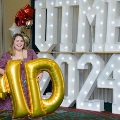 Individual standing indoors holding large gold “M” and “D” balloons in front of illuminated “UTMB 24” letters, with red, white, gold, and silver balloon clusters arranged alongside a curtain backdrop.