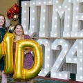 Two individuals stand indoors holding large gold “M” and “D” balloons in front of illuminated “UTMB 24” letters, with red, white, gold, and silver balloon clusters arranged on both sides against a curtain backdrop.