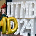 Individual standing indoors holding large gold “M” and “D” balloons in front of illuminated “UTMB 24” letters, with red, white, gold, and silver balloon clusters arranged on both sides against a curtain backdrop. Provide your feedback on BizChat