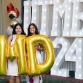 Two individuals stand indoors holding large gold “M” and “D” balloons in front of illuminated “UTMB 24” letters, with red, white, gold, and silver balloon clusters arranged on both sides against a curtain backdrop.