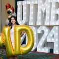 Individual standing indoors holding large gold “M” and “D” balloons in front of illuminated “UTMB 24” letters, with red, gold, silver, and white balloon clusters arranged beside a curtain backdrop.