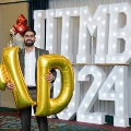 Individual standing indoors holding large gold “M” and “D” balloons in front of illuminated “UTMB 24” letters, with red, white, gold, and silver balloon clusters on both sides against a blue curtain backdrop.