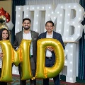 Three individuals stand indoors holding large gold “MD” balloons in front of illuminated “UTMB 24” letters, with red, white, gold, and silver balloon clusters on both sides against a curtain backdrop.