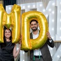 Two individuals stand indoors holding large gold “M” and “D” balloons in front of illuminated “UTMB 24” letters, with decorative balloon arrangements on both sides against a curtain backdrop.
