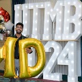 Individual standing indoors holding large gold “MD” balloons in front of illuminated “UTMB 24” letters, with red, white, gold, and silver balloons arranged on both sides against a blue curtain backdrop.