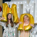 Two individuals pose indoors holding large gold “M” and “D” balloons in front of illuminated “UTMB 24” letters, with decorative red, white, gold, and silver balloon bundles on both sides against a curtain backdrop.