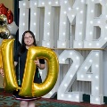 Individual poses with large gold “MD” balloons in front of illuminated “UTMB 2024” display with red, white, and gold decorations at an Alpha Omega Alpha Texas Alpha Chapter event.