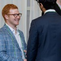 Individual wearing green honor cords and glasses speaks with another attendee in a suit during an indoor Alpha Omega Alpha Texas Alpha Chapter event at UTMB.