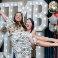 Two individuals pose in front of illuminated “UTMB 2024” display, with one lifting the other while holding them horizontally, with red, white, and gold balloon decorations at an Alpha Omega Alpha Texas Alpha Chapter event.