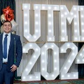 Individual in a blue suit with green honor cords stands in front of illuminated “UTMB 2024” display with red, white, and gold balloon decorations at an Alpha Omega Alpha Texas Alpha Chapter event.