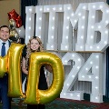 Two individuals stand in front of illuminated “UTMB 2024” display holding large gold “M” and “D” balloons with red, white, and gold decorations at an Alpha Omega Alpha Texas Alpha Chapter event.