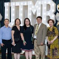 Seven individuals stand together in front of illuminated “UTMB 2024” letters with red, white, and gold balloon decorations at an Alpha Omega Alpha Texas Alpha Chapter event.