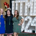 Three individuals stand together in front of illuminated “UTMB 2024” display, two wearing green honor cords, with red, white, and gold balloon decorations at an Alpha Omega Alpha Texas Alpha Chapter event.