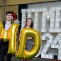 Two individuals stand in front of illuminated “UTMB 2024” display holding large gold “M” and “D” balloons with red, white, and gold decorations at an Alpha Omega Alpha Texas Alpha Chapter event.