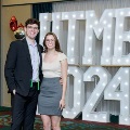 Two individuals stand together in front of illuminated “UTMB 2024” display, one wearing green honor cords, with red, white, and gold balloon decorations at an Alpha Omega Alpha Texas Alpha Chapter event.