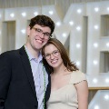Two individuals stand close together in front of illuminated “UTMB” letters, one wearing green honor cords, at an Alpha Omega Alpha Texas Alpha Chapter event.