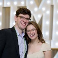 Two individuals stand close together in front of illuminated “UTMB” letters, one wearing green honor cords, at an Alpha Omega Alpha Texas Alpha Chapter event.