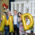 Three individuals pose in front of illuminated “UTMB” letters holding large gold “M” and “D” balloons, with red, white, and gold decorations at an Alpha Omega Alpha Texas Alpha Chapter event.