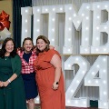 Three individuals pose together in front of illuminated “UTMB 2024” display with red, white, and gold balloon decorations at an Alpha Omega Alpha Texas Alpha Chapter event.