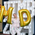 Individual holding large gold “M” and “D” balloons in front of illuminated “UTMB 2024” display with red, white, and gold balloon decorations at an Alpha Omega Alpha Texas Alpha Chapter event.