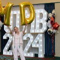 Individual raises large gold “M” and “D” balloons overhead in front of illuminated “UTMB 2024” display with red, white, and gold balloon decorations at an Alpha Omega Alpha Texas Alpha Chapter event.