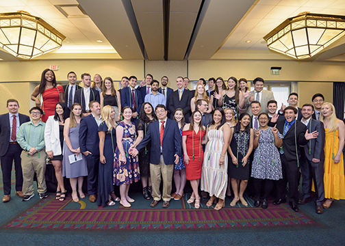 Approximately 35 to 45 attendees stand and sit together posing for a large group photo in a ballroom with chandeliers and patterned carpet during the Alpha Omega Alpha Texas Chapter UTMB 2022 Fall Mixer.
