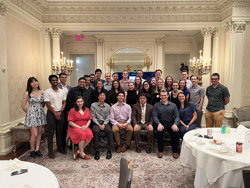 Large group of attendees posed together in an ornate indoor venue with decorative walls and chandeliers, seated and standing in rows during the Alpha Omega Alpha Texas Chapter UTMB 2022 Fall Mixer event; tables with drinks visible in foreground.
