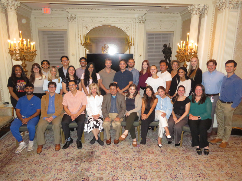 Large group of UTMB Alpha Omega Alpha members posing for a formal photo in an ornate room with chandeliers, decorative molding, and a patterned carpet, arranged in seated and standing rows.
