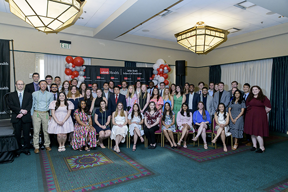 A large group of inductees and guests pose together at the Alpha Omega Alpha 2023 Induction Ceremony at UTMB, arranged in rows in a ballroom with chandeliers and a UTMB Health backdrop.