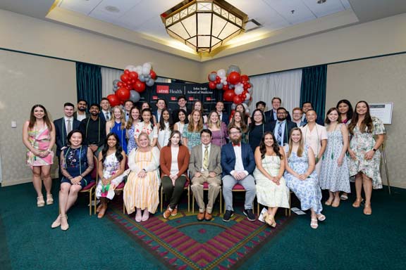 Large group of individuals pose indoors in front of a UTMB Health and John Sealy School of Medicine backdrop, framed by red, white, and gray balloon arches, with some seated in front and others standing behind in a well-lit event space.