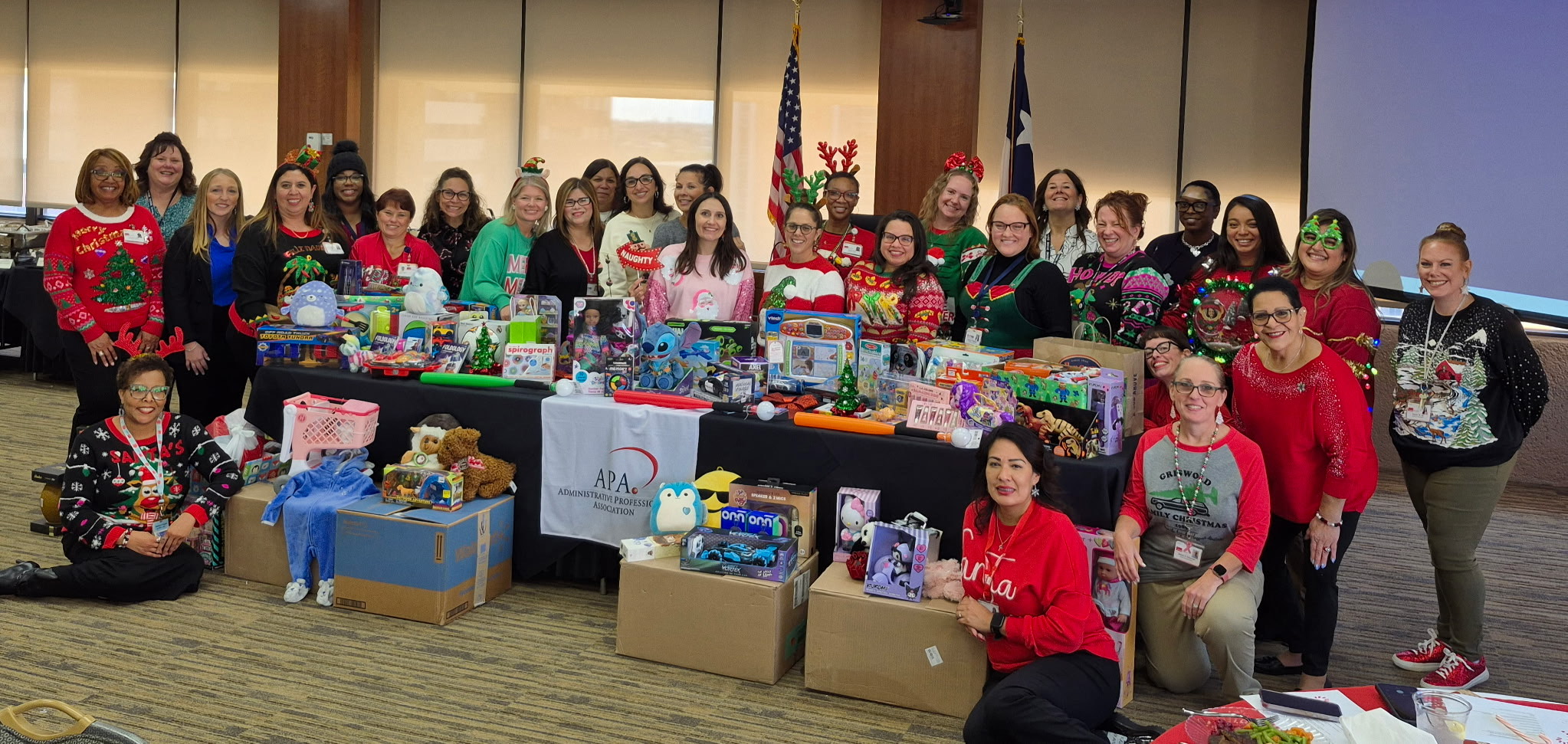 Group of Administrative Professionals Association members at UTMB standing behind tables filled with donated toys during a holiday toy drive event.