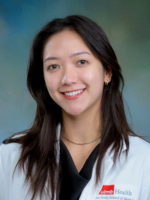 Professional headshot of Elizabeth Denney with medium length dark brown hair, wearing a black top under a whitecoat