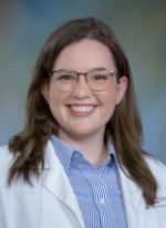 Professional headshot of Gracie Geddie with medium length highlighted brown hair, wearing a blue top under a whitecoat