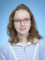 Professional headshot of Katherine Lomoc with medium length brown hair, wearing a blue striped shirt in front of a blue background