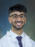 Professional headshot of Yasein Shamsulhooda with short black hair, wearing a blue tie under a whitecoat