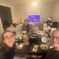 Group seated around a dining table with a shared meal, including multiple dishes, bowls, and drinks, in a warmly lit room with a television and small decorative tree in the background during a fellowship gathering.
