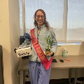 Individual in scrubs wearing a red “Doctor of the Day” sash holds a small trophy and a decorated gift box, standing in a clinical office space with a desk and window in the background. Provide your feedback on BizChat