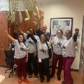 Group of fellowship participants standing in a clinical room with arms raised, wearing matching shirts, surrounded by festive decorations and a large gold display hanging behind them.
