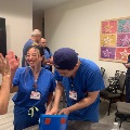 Group of individuals in scrubs gathered around a table participating in a hands-on team activity with a colorful box, inside a conference room decorated with wall art.