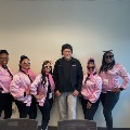 Group of individuals posing indoors wearing coordinated pink jackets and dark sunglasses alongside another individual in a black jacket, standing in front of a neutral wall.