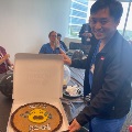 Individual in scrubs holding an open cake box with a decorated cookie cake on a table, while others gather in a conference room during a UTMB fellowship celebration.